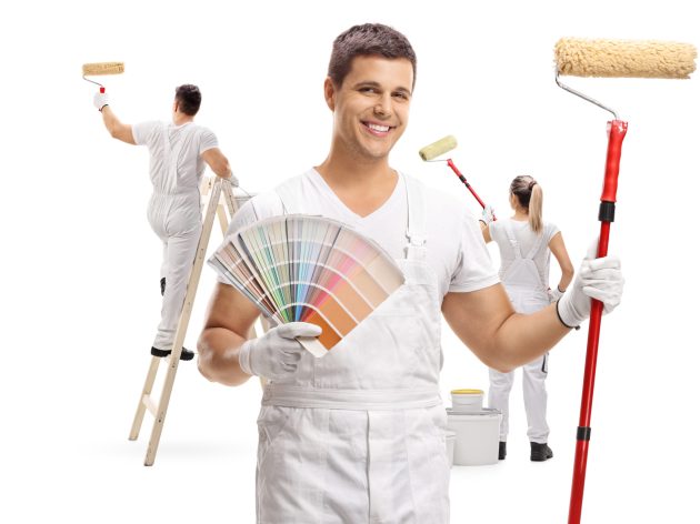 Painter holding a color swatch and a paint roller with two painters painting behind him isolated on white background