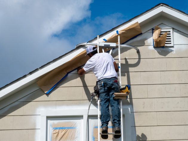 Painter on a ladder masking off trim at the peak of a house befo
