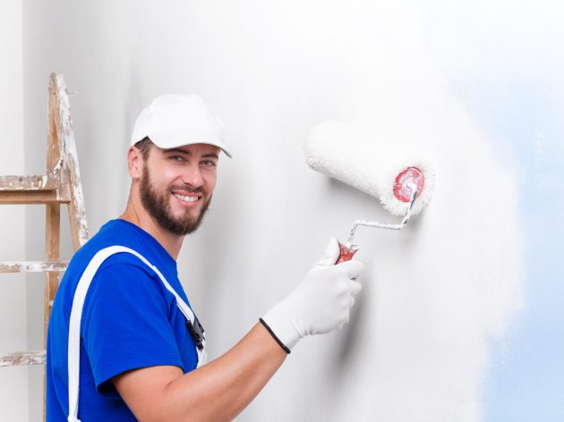 Portrait of handsome young smiling painter in white dungarees, blue t-shirt, cap and gloves painting a wall with paint roller, looking at camera